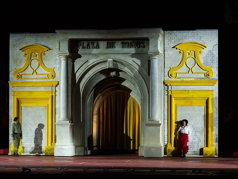 Szene: Kulisse mit einem „Plaza de Toros“-Tor in Gelb und Weiß, einer Frau in rotem Rock und einem Soldaten in Uniform