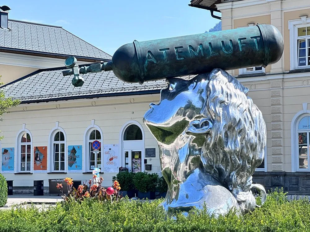 glänzende Skulptur einer Frau ab den Schultern, mit einer Sauerstoffflasche mit der Aufschrift "Atemluft" zentriert auf ihrem Kopf platziert, vor einem Gebäude mit Hecken und Blumen, gekennzeichnet durch ein Schild "Atemluft 2024 Xenia Hausner". Die Skulptur hat eine Höhe von etwa drei Metern und besteht aus poliertem Aluminium und Bronze.