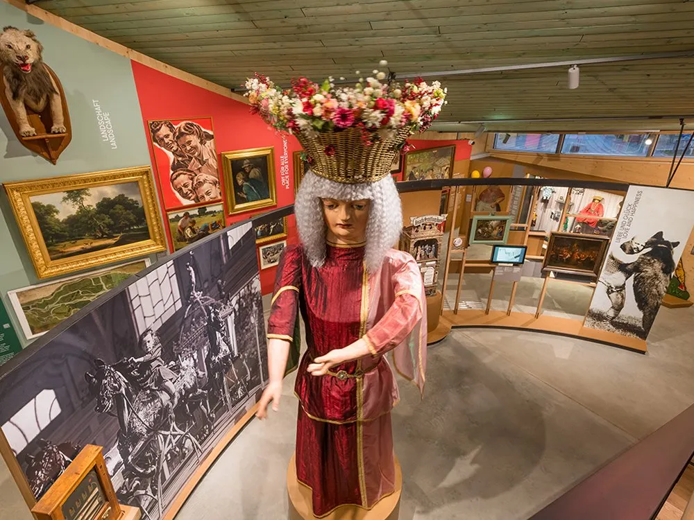 Fortuna-Figur mit Blumenkorb-Krone, umgeben von historischen Bildern und Exponaten, darunter ein Pferdekarussell-Foto.