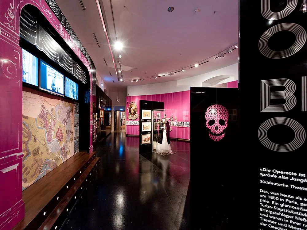Ausstellungsraum: Historische Karte, Bildschirme, Kunst und Kleid in Vitrine, rosa Wände mit Schädel-Design und Operetten-Texten
