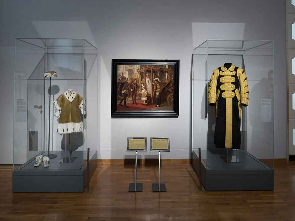 Ausstellungsraum: Vitrinen mit goldenem Gewand und Schuhen, gelb-schwarzem Uniformmantel, Gemälde einer königlichen Kutsche