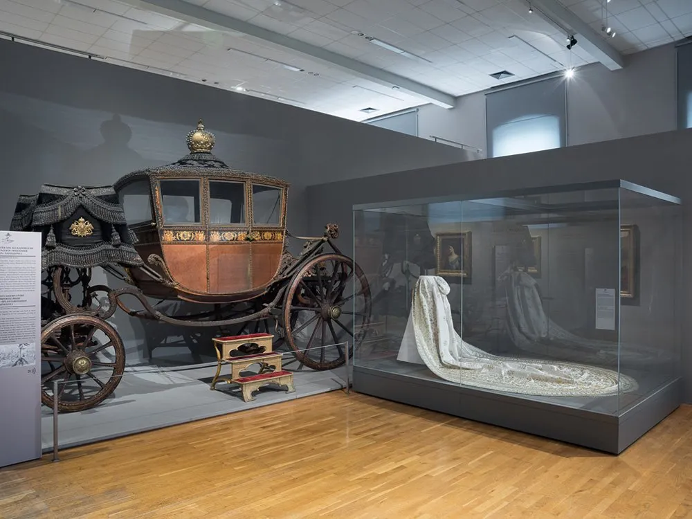 Ausstellungsraum: Historische Kutsche mit goldenen Verzierungen und Stufen neben einer Vitrine mit einem weißen, bestickten Kleid