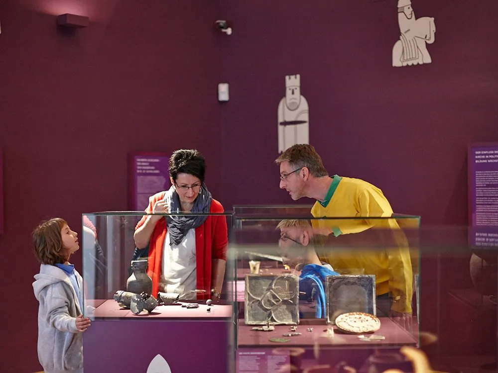 Ausstellungsraum mit Vitrinen, die Artefakte und ein Modell eines antiken Marktes zeigen, sowie einer beleuchteten Säule im Hintergrund