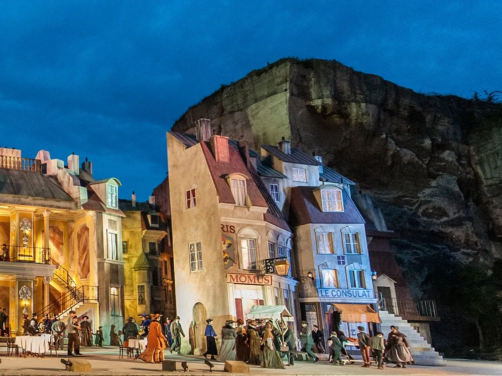 Kulisse eines pariserischen Stadtteils mit beleuchteten Gebäuden wie Café Momus und Le Consulat, Figuren in historischer Kleidung