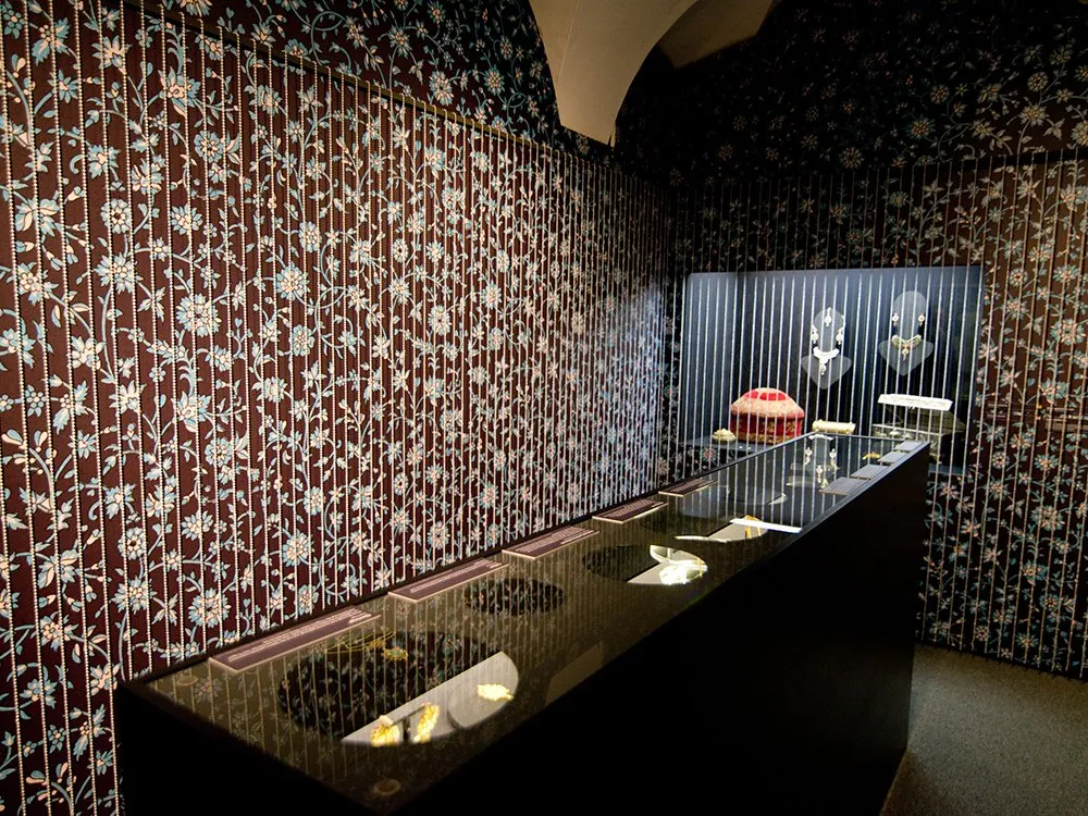 Ausstellungsraum mit dunkelbraunen, floral gemusterten Wänden, einer Vitrine mit kleinen Kunstwerken