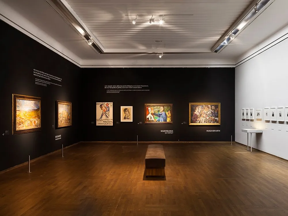 Ausstellungsraum: Zwei schwarze und eine weiße Wand, mit Gemälden, Bank zentral im Raum 