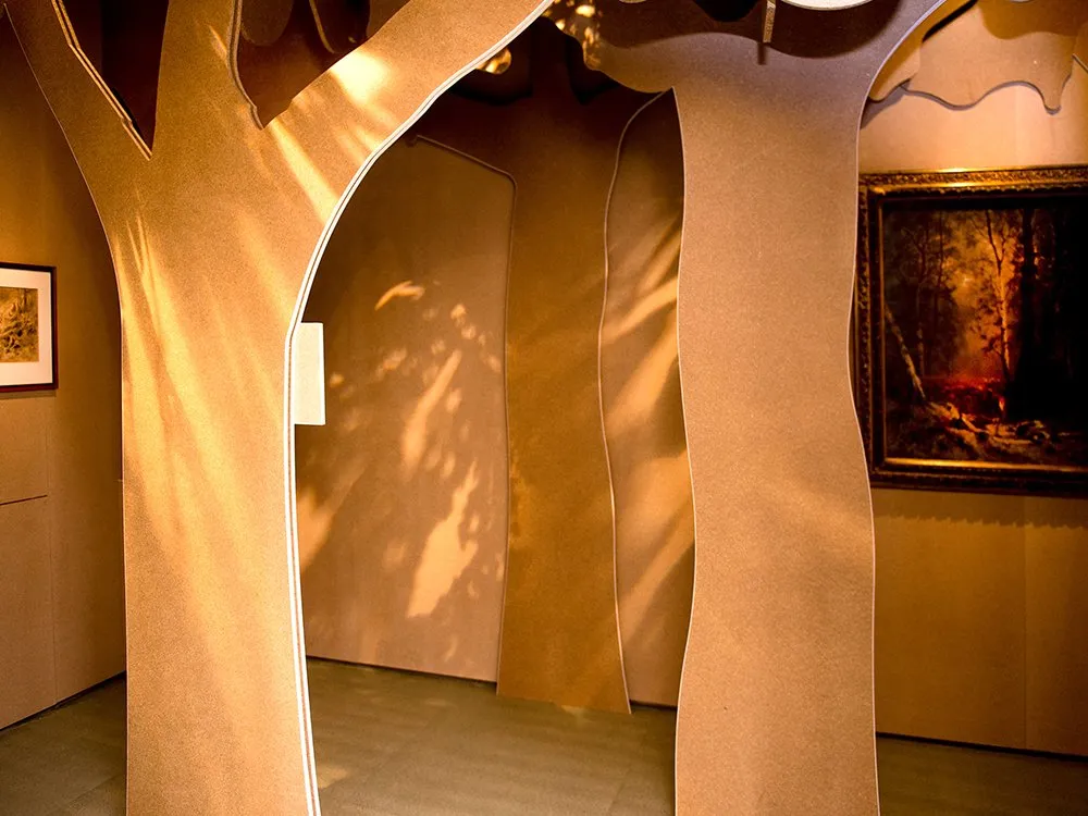 Ausstellungsraum: Ein Raum mit baumförmigen, braunen Wänden, durch die Lichtschatten fallen, umgeben von Gemälden, darunter eines mit Waldszene
